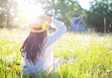 achterkant van vrouw met hoed op, zittend in een zonovergoten grasveld, kijkend in de verte.