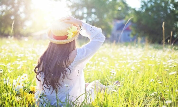 achterkant van vrouw met hoed op, zittend in een zonovergoten grasveld, kijkend in de verte.